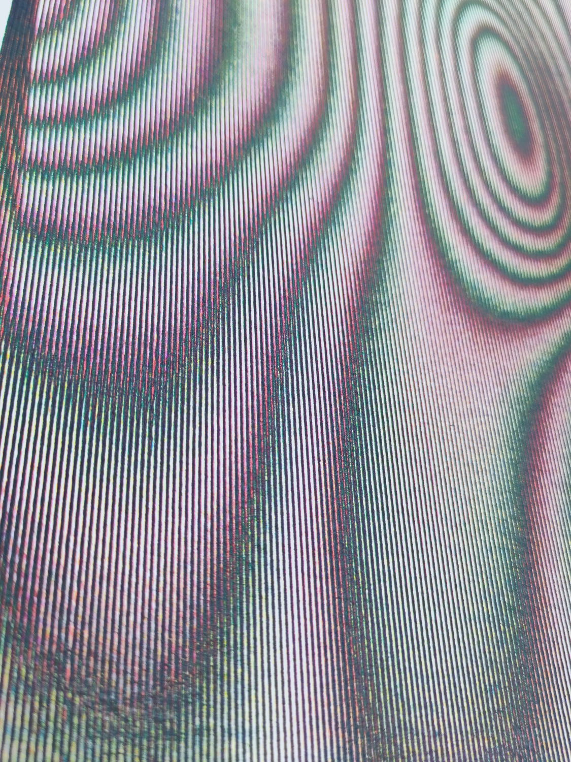 A close-up view of an abstract pattern that creates a moiré effect. The artwork consists of fine, closely spaced lines that are slightly curved, producing an illusion of waves and ripples. The colors in the image are predominantly shades of green and pink, which blend together to create a sense of depth and movement.