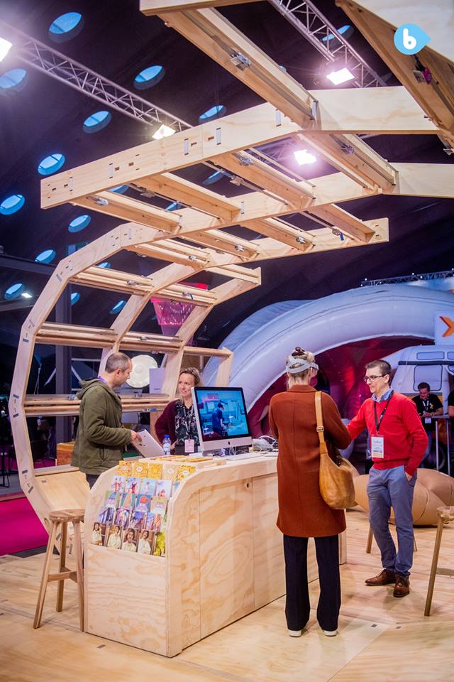 Inside the structure, looking at the central counter with sample flyers, computers, and people chatting. Above the open woodwork, held together with tension belts, crosses diagonally to the other side where it comes down.